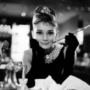 Optimized by JPEGmini 3.14.12.71901706 0x4a57e0e9 Portrait culte en noir et blanc d'Audrey Hepburn (Holly Golightly) accoudée à une table, portant un collier de perles, un diadème et tenant son long porte-cigarette.