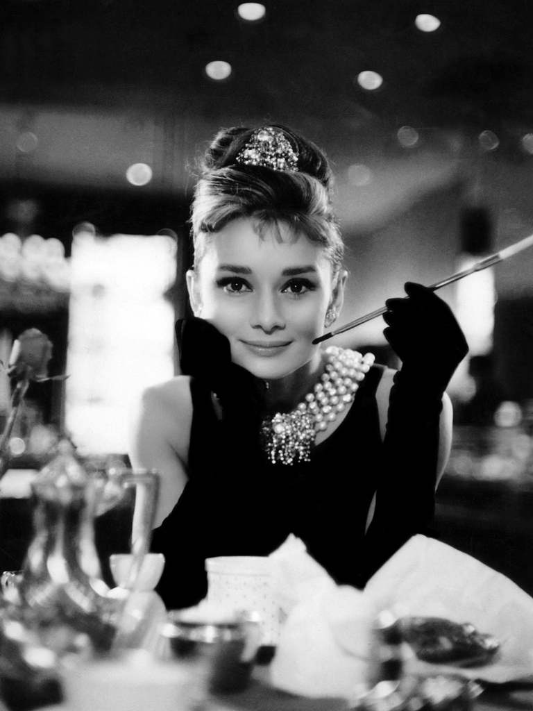 Optimized by JPEGmini 3.14.12.71901706 0x4a57e0e9 Portrait culte en noir et blanc d'Audrey Hepburn (Holly Golightly) accoudée à une table, portant un collier de perles, un diadème et tenant son long porte-cigarette.