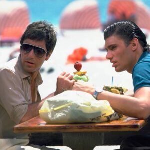 Al Pacino (Tony Montana) et Steven Bauer (Manny) assis à une table de plage à Miami avec des cocktails, scène du film Scarface, ambiance années 80 colorée.