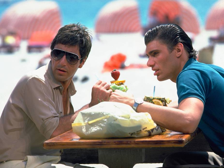 BRAP103 - 4058415 Al Pacino (Tony Montana) et Steven Bauer (Manny) assis à une table de plage à Miami avec des cocktails, scène du film Scarface, ambiance années 80 colorée.