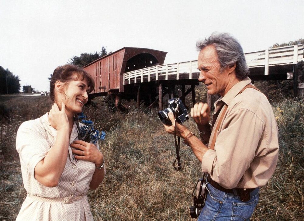 Photo couleur de Meryl Streep et Clint Eastwood dans le film Sur la route de Madison. Elle sourit avec des fleurs bleues à la main tandis qu'il s'apprête à la photographier devant un pont couvert en bois.