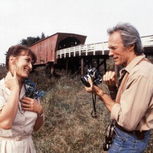 BRCL100 - 1707408 Photo couleur de Meryl Streep et Clint Eastwood dans le film Sur la route de Madison. Elle sourit avec des fleurs bleues à la main tandis qu'il s'apprête à la photographier devant un pont couvert en bois.