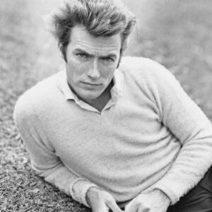 Portrait en noir et blanc d'un jeune Clint Eastwood allongé sur l'herbe, portant un pull clair sur une chemise à col, fixant l'objectif avec intensité.