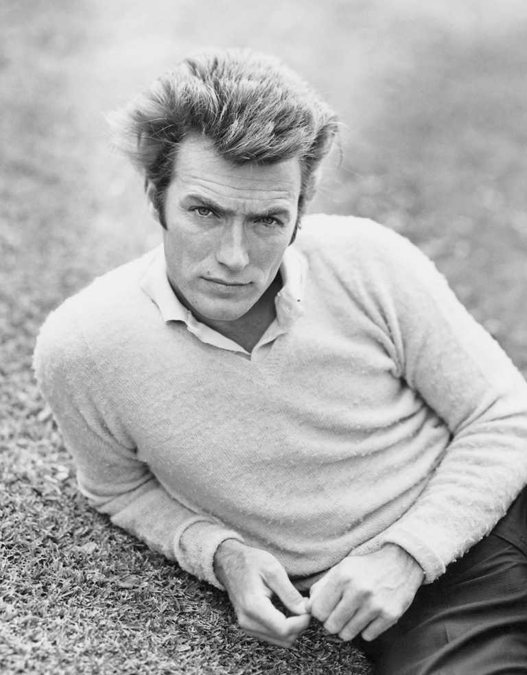 BRCL111 BAL_4062972 Portrait en noir et blanc d'un jeune Clint Eastwood allongé sur l'herbe, portant un pull clair sur une chemise à col, fixant l'objectif avec intensité.