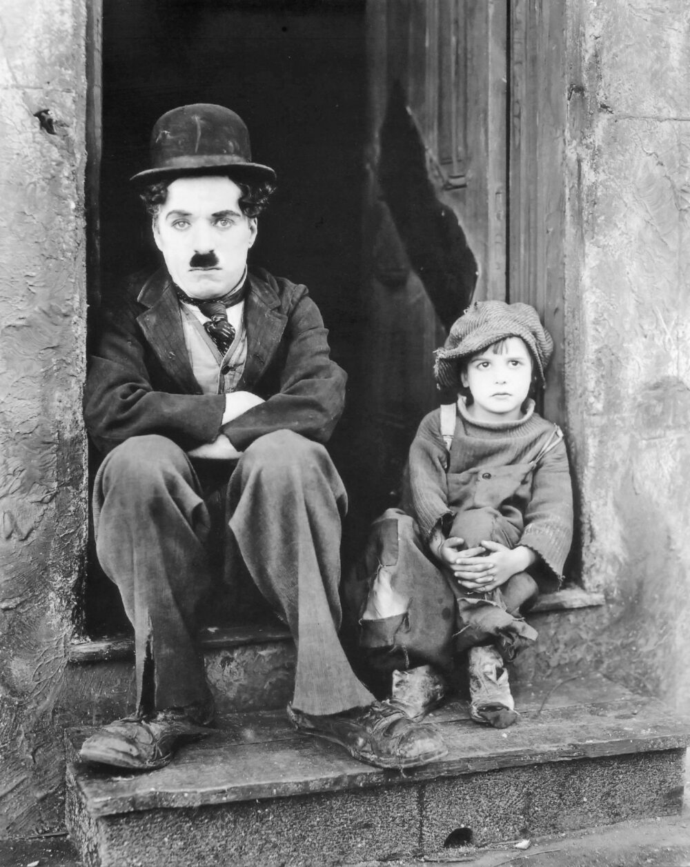 CC06 OK Photographie iconique en noir et blanc de Charlie Chaplin et du jeune acteur Jackie Coogan assis côte à côte sur un perron, dans leurs costumes fidèles au film Le Kid.