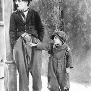 Photographie de plateau en noir et blanc de Charlie Chaplin (Charlot) s'appuyant contre un poteau, tandis que son jeune partenaire pointe du doigt vers la gauche, tous deux vêtus de leurs costumes fidèles au film Le Kid.