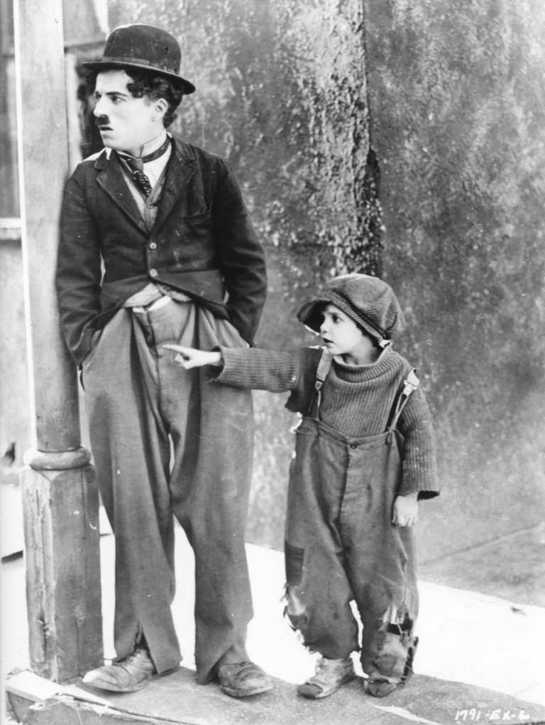 Photographie de plateau en noir et blanc de Charlie Chaplin (Charlot) s'appuyant contre un poteau, tandis que son jeune partenaire pointe du doigt vers la gauche, tous deux vêtus de leurs costumes fidèles au film Le Kid.