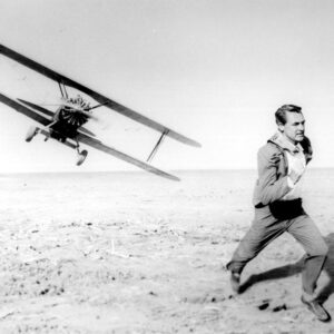MBDNOBY EC004 Photographie de plateau en noir et blanc du film La Mort aux trousses (1959) montrant Cary Grant en costume, courant dans un champ désertique alors qu'un avion biplan le poursuit à basse altitude en arrière-plan.