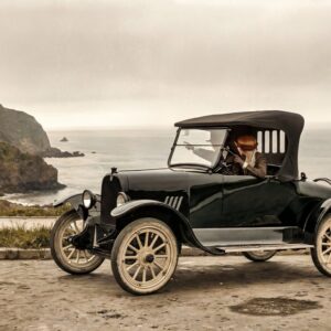 L'Échappée Belle (1919) : Affiche Auto Rétro | Fuzars Photographie historique colorisée de 1919 montrant un couple en voiture ancienne garée sur une route à flanc de falaise, admirant l'océan.