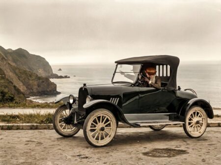 Photographie historique colorisée de 1919 montrant un couple en voiture ancienne garée sur une route à flanc de falaise, admirant l'océan.