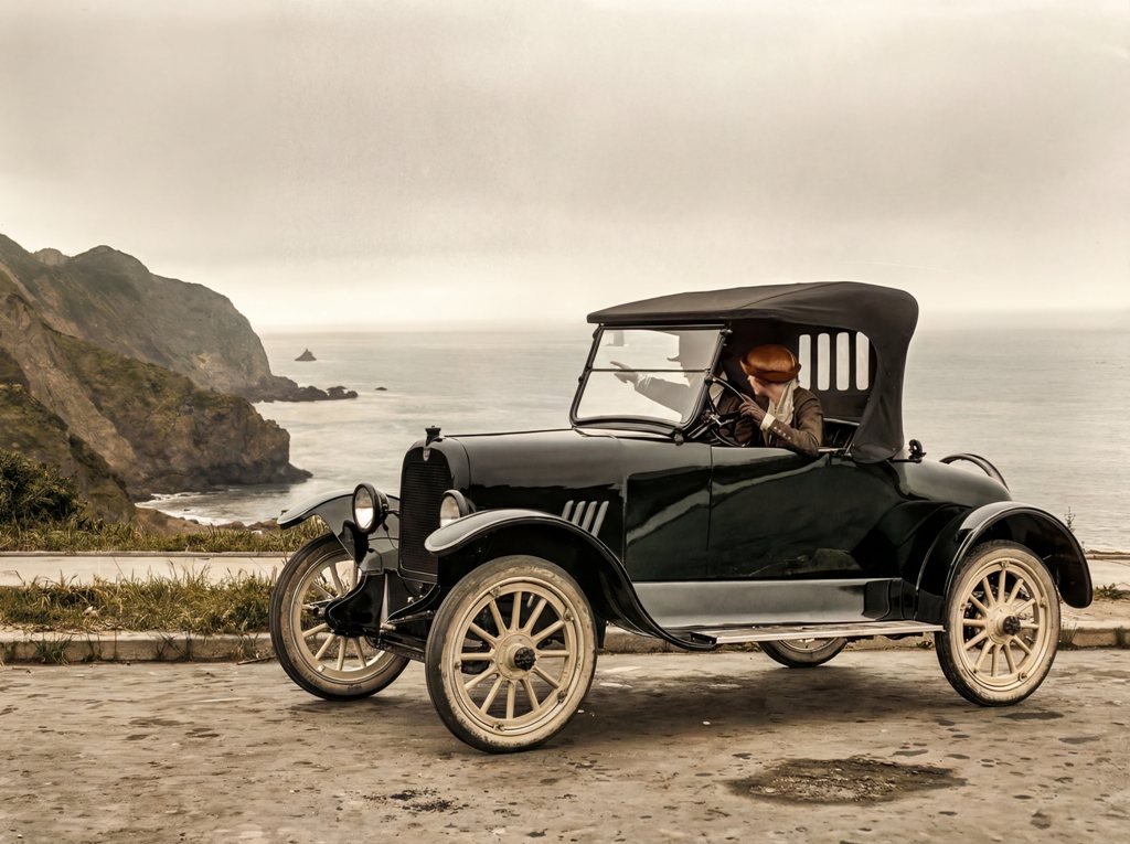 L'Échappée Belle (1919) : Affiche Auto Rétro | Fuzars Photographie historique colorisée de 1919 montrant un couple en voiture ancienne garée sur une route à flanc de falaise, admirant l'océan.