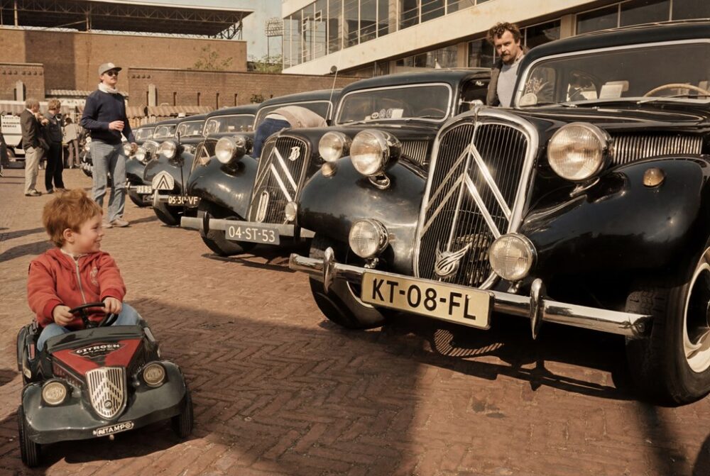 Citroën Traction Avant Paris-Moscou 1984 Affiche publicitaire vintage colorisée de 1984 montrant la Citroën Traction Avant n°116 en pleine course sur une piste de terre boueuse en Russie, acclamée par la foule, sous un ciel nuageux. Titres "PARIS-MOSCOU '84" et "CITROËN TRACTION AVANT".