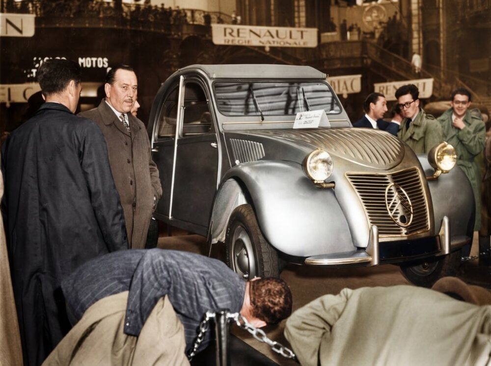Photographie historique colorisée montrant la présentation de la première Citroën 2CV au Salon de l'Auto de Paris en 1948, entourée d'une foule de curieux.