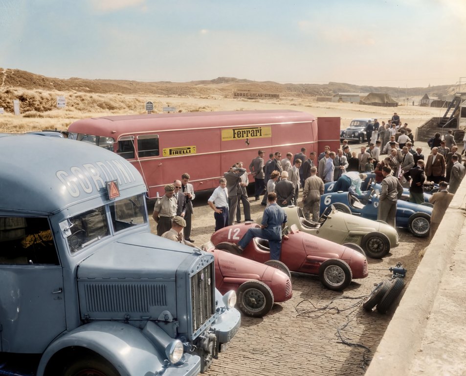 Grand Prix 1952 : Effervescence des Stands | Fuzars Photographie historique colorisée en vue plongée depuis une tribune, montrant quatre monoplaces de course de 1952 (dont une Ferrari rouge à l'arrière, une bleue, une grise et une verte) préparées par leurs mécaniciens dans la voie des stands, entourées d'une foule en costume.