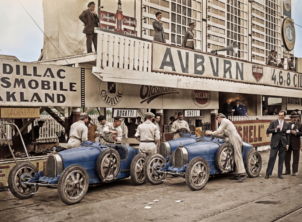 Bugatti au Grand Prix 1926 : Affiche Auto Vintage | Fuzars Photographie historique colorisée du Grand Prix d'Espagne 1926, montrant des mécaniciens préparant deux légendaires Bugatti bleues dans la voie des stands, sous des panneaux publicitaires d'époque.