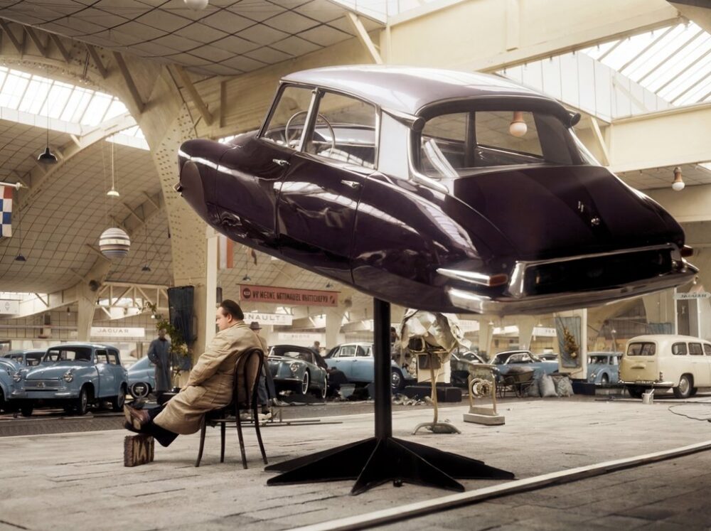 La DS Volante (1958) : Ovni Rétro-Futuriste | Fuzars Photographie historique colorisée d'un salon automobile en 1958 montrant la carrosserie d'une Citroën DS aubergine exposée en lévitation sur un pied, avec un homme assis calmement en dessous.