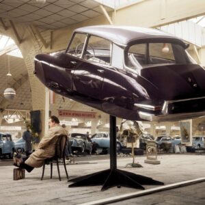 Photographie historique colorisée d'un salon automobile en 1958 montrant la carrosserie d'une Citroën DS aubergine exposée en lévitation sur un pied, avec un homme assis calmement en dessous.
