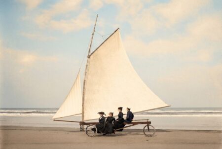 Photographie historique colorisée d'un char à voile ancien sur une plage immense. Quatre passagers élégants en costumes de la Belle Époque sont installés sous la grande voile blanche.