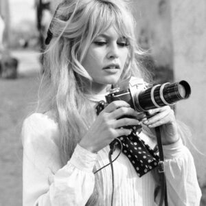 Photographie en noir et blanc de Brigitte Bardot portant une blouse blanche et un foulard à pois, tenant un appareil photo professionnel avec un long téléobjectif.