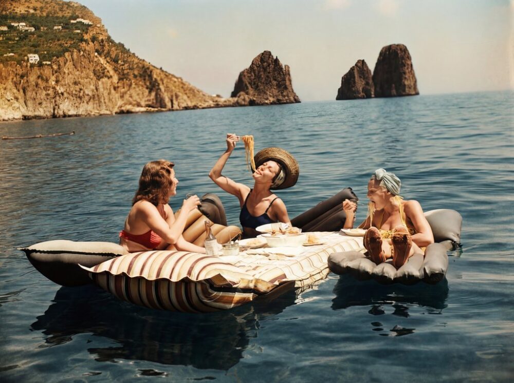 La Dolce Vita : Pique-nique à Capri | Fuzars Photographie vintage colorisée montrant trois femmes riant et mangeant des spaghettis sur des matelas gonflables flottant sur la mer à Capri.