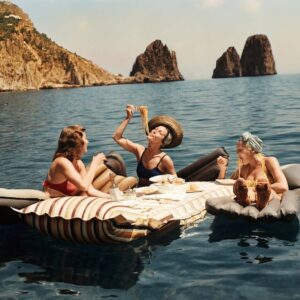 La Dolce Vita : Pique-nique à Capri | Fuzars Photographie vintage colorisée montrant trois femmes riant et mangeant des spaghettis sur des matelas gonflables flottant sur la mer à Capri.