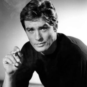 Portrait photographique vintage en noir et blanc de l'acteur français Alain Delon jeune, fixant l'objectif avec un regard intense et tenant une cigarette allumée.