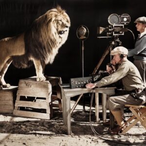 Photographie historique colorisée de 1928 montrant un lion rugissant devant un microphone et une caméra de cinéma, entouré de techniciens en studio.