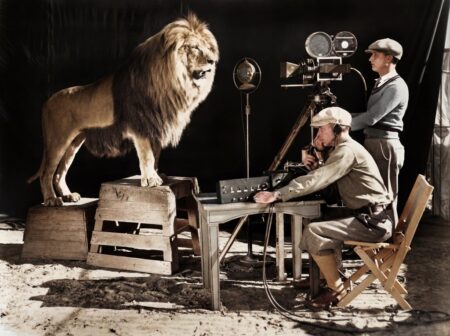 Photographie historique colorisée de 1928 montrant un lion rugissant devant un microphone et une caméra de cinéma, entouré de techniciens en studio.