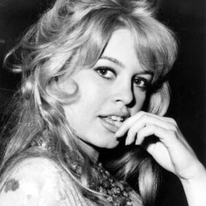 BRBB102 - 1927106 Photographie en noir et blanc de Brigitte Bardot en 1959 en plan rapproché. Elle pose avec sa célèbre coiffure volumineuse, le regard tourné vers l'objectif, la main délicatement posée près de sa bouche entrouverte, portant un imposant collier de perles et un haut à motifs.