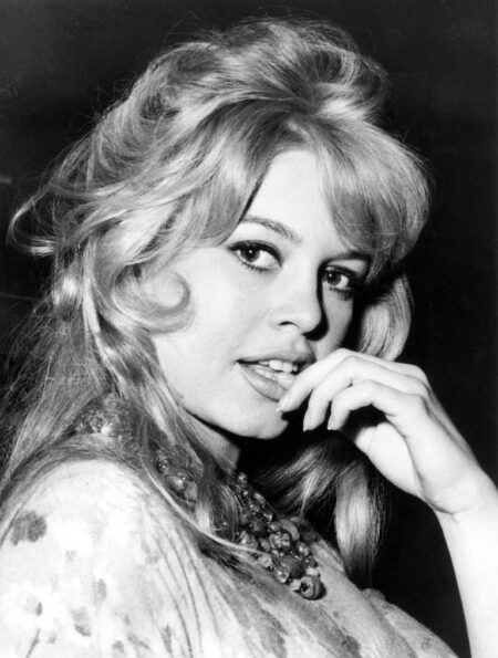 Photographie en noir et blanc de Brigitte Bardot en 1959 en plan rapproché. Elle pose avec sa célèbre coiffure volumineuse, le regard tourné vers l'objectif, la main délicatement posée près de sa bouche entrouverte, portant un imposant collier de perles et un haut à motifs.
