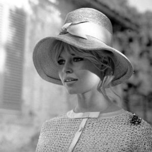 Portrait lumineux en noir et blanc de Brigitte Bardot en 1961, portant un grand chapeau clair avec un ruban, une tenue texturée et une broche, le visage partiellement ombragé.