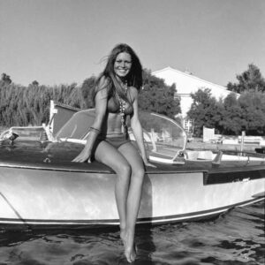 Photographie artistique en noir et blanc de Brigitte Bardot souriante, assise sur le bord d'un bateau devant sa villa La Madrague à Saint-Tropez en 1973.