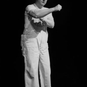 Photographie en pied et en noir et blanc de Coluche sur scène en 1977, portant sa célèbre salopette à rayures, un t-shirt blanc et des lunettes rondes, effectuant un geste humoristique sur un fond noir profond.