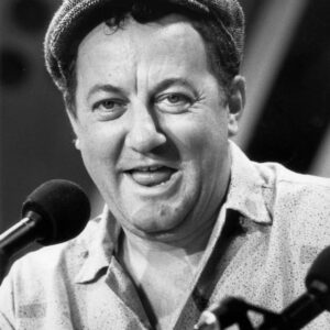 Photographie en noir et blanc de l'humoriste français Coluche en 1985, souriant face à des micros et portant sa célèbre casquette gavroche en tweed.