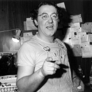 Photo noir et blanc de Coluche en 1975, portant des lunettes rondes et une salopette rayée, pointant du doigt vers l'appareil photo depuis sa loge avec des feuillets épinglés au mur et une mallette de maquillage éclairée.