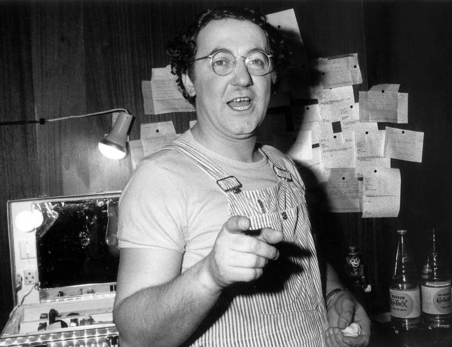 Photo noir et blanc de Coluche en 1975, portant des lunettes rondes et une salopette rayée, pointant du doigt vers l'appareil photo depuis sa loge avec des feuillets épinglés au mur et une mallette de maquillage éclairée.