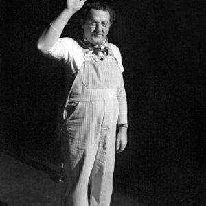 Photographie en noir et blanc de Coluche sur scène en 1981, vêtu de sa salopette à rayures et d'un foulard, levant la main pour saluer le public avec une table visible au premier plan.