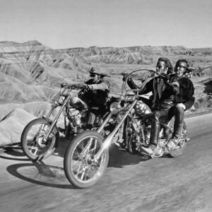 Photographie en noir et blanc du film Easy Rider (1969) montrant Dennis Hopper, Peter Fonda et Jack Nicholson sur des motos choppers. Ils roulent sur une route dégagée au milieu d'un paysage de canyons désertiques. Jack Nicholson est assis à l'arrière de la moto de Peter Fonda.