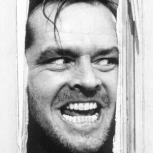 Gros plan en noir et blanc de Jack Nicholson (Jack Torrance) dans le film The Shining. Il passe son visage à travers la fente d'une porte en bois fracassée, affichant un sourire dément et un regard maniaque.