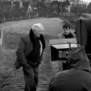 Photographie en noir et blanc sur le tournage du film Un Singe en Hiver, montrant Jean Gabin et Jean-Paul Belmondo debout sur des marches en extérieur avec un village côtier en arrière-plan, face à une caméra de cinéma et un membre de l'équipe.