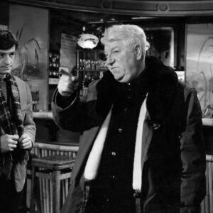 Photographie en noir et blanc du film Un Singe en Hiver avec Jean Gabin en manteau épais pointant le doigt, observé par Jean-Paul Belmondo dans un décor de bar avec une barmaid en arrière-plan.