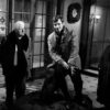 Affiche noir et blanc du film Un Singe en Hiver montrant Jean Gabin de face, observant Jean-Paul Belmondo qui est assis au-dessus de lui sur le dossier d'un gros pouf en cuir capitonné, dans un grand hall avec des portes vitrées. Une femme en robe de chambre sombre les regarde depuis l'arrière-plan à droite.