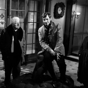Affiche noir et blanc du film Un Singe en Hiver montrant Jean Gabin de face, observant Jean-Paul Belmondo qui est assis au-dessus de lui sur le dossier d'un gros pouf en cuir capitonné, dans un grand hall avec des portes vitrées. Une femme en robe de chambre sombre les regarde depuis l'arrière-plan à droite.