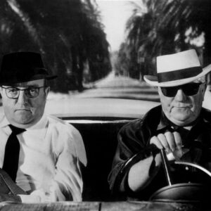 Photographie en noir et blanc du film Le Cave se rebiffe montrant Bernard Blier avec un chapeau noir et des lunettes de vue, assis côté passager à côté de Jean Gabin qui conduit une décapotable en portant un chapeau blanc et des lunettes de soleil, sur une route bordée de palmiers.