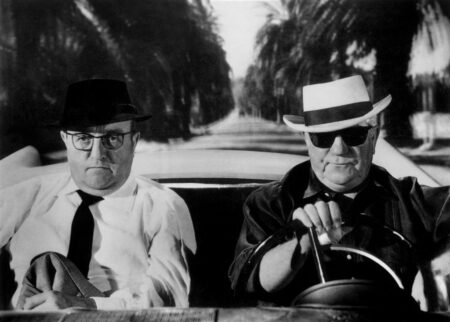 Photographie en noir et blanc du film Le Cave se rebiffe montrant Bernard Blier avec un chapeau noir et des lunettes de vue, assis côté passager à côté de Jean Gabin qui conduit une décapotable en portant un chapeau blanc et des lunettes de soleil, sur une route bordée de palmiers.