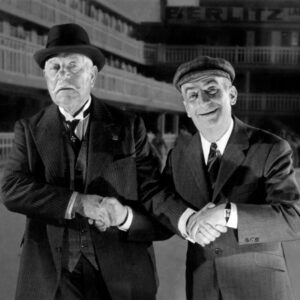Photographie en noir et blanc de Jean Gabin en costume et chapeau serrant la main de Louis de Funès souriant en casquette, issue du film Le Tatoué.