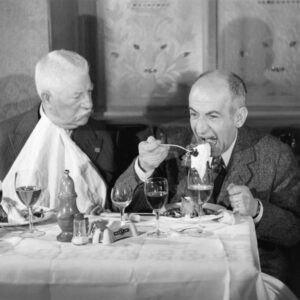 Photographie en couleur du film Le Tatoué montrant Jean Gabin, l'air impassible avec une serviette nouée au cou, observant Louis de Funès qui mange goulûment à une table de restaurant dressée avec des verres de vin.