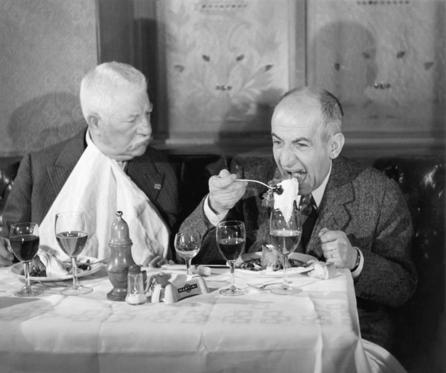 BRLDF118NB - 1780831 Photographie en couleur du film Le Tatoué montrant Jean Gabin, l'air impassible avec une serviette nouée au cou, observant Louis de Funès qui mange goulûment à une table de restaurant dressée avec des verres de vin.