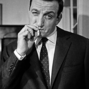 Portrait en noir et blanc de Lino Ventura dans le rôle de Fernand Naudin. Il est de face, vêtu d'un costume sombre et d'une cravate, avec une expression sérieuse et déterminée.