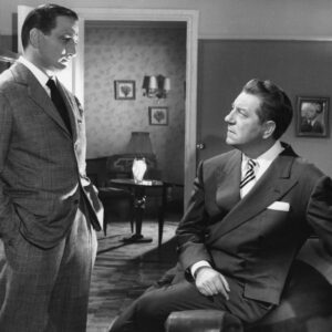 Photographie noir et blanc du film Touchez pas au grisbi : Jean Gabin assis sur un canapé regarde Lino Ventura debout, tous deux en costume élégant dans un décor de salon d'époque.